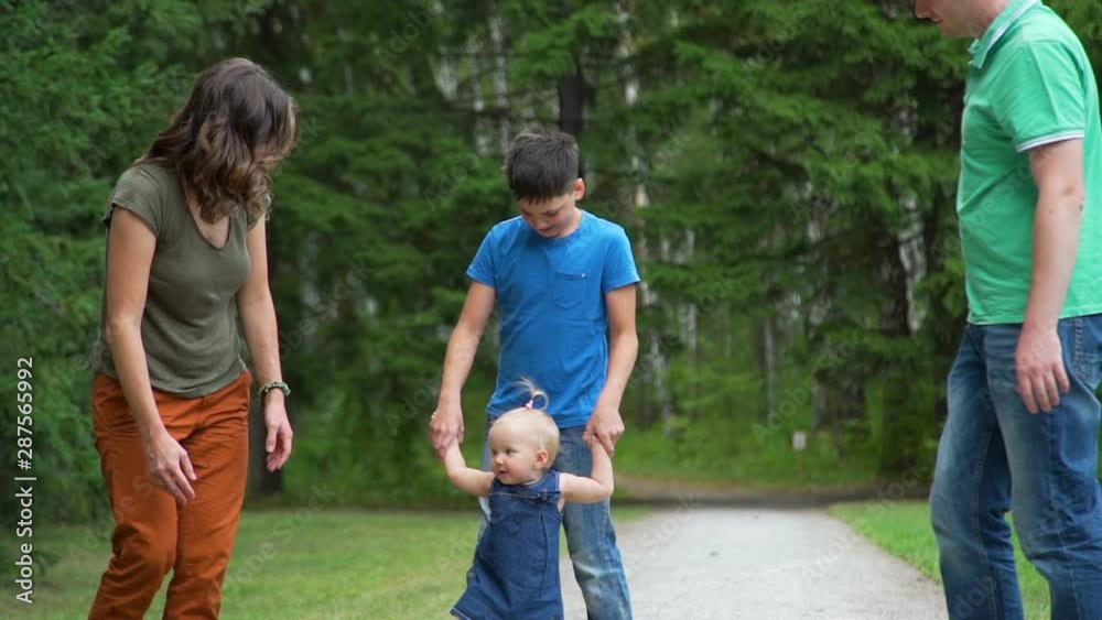 Cute Baby Girl Walking with Parents and Brother Outdoors. Child Making First Steps. Happy Childhood and Child Development Concept Cute Baby Girl Walking with Parents and Brother Outdoors. Child Making First Steps. Happy Childhood and Child Development Concept