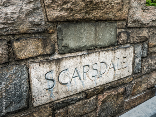 A sign reading Scarsdale with letters engraved in limestone in a stone wall along a stair leading to the Bronx River walkway trail.