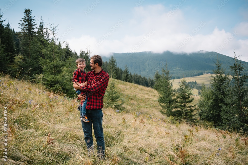Happy father with his young son sitting on the grass on a background of green forest, mountains and sky with clouds. Friendship concept. Father with his son hiking. Father's Day
