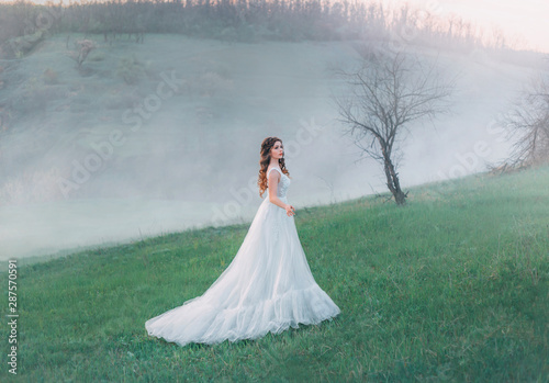 A fabulous woman stands against the backdrop of a mountain landscape in a white vintage ball gown. Tale of Cinderella with a doll face. Landscape hills in the cold autumn. Fog and haze at dusk. 