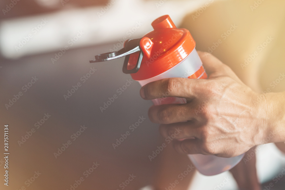 Naklejka premium Close up of man's hand holding a classic fitness shaker with pre-workout drink in it. Sports nutrition concept. Horizontal shot