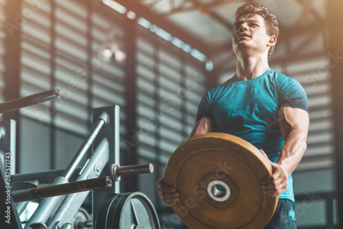 Portrait of athletic man with the muscular body holding a barbell disk while having a workout at gym. Horizontal shot