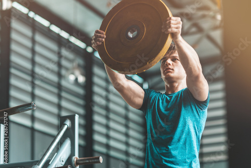 Portrait of athletic man with the muscular body holding a barbell disk while having a workout at gym. Horizontal shot