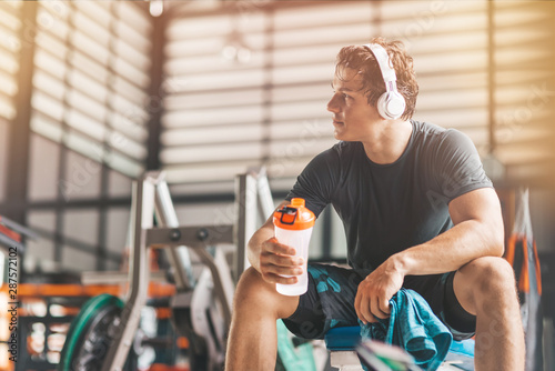 Portait of athletic man in headphones looking aside while listening to music and holding a towel and a classic fitness shaker with pre-workout drink in it. Horizontal shot
