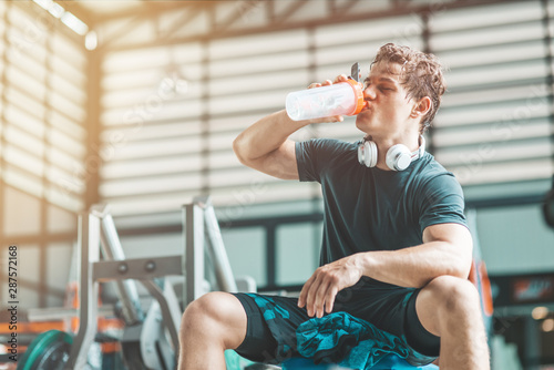 Portait of tired athletic man relaxing and drinking pre-workout drink from a classic fitness shaker. Horizontal shot