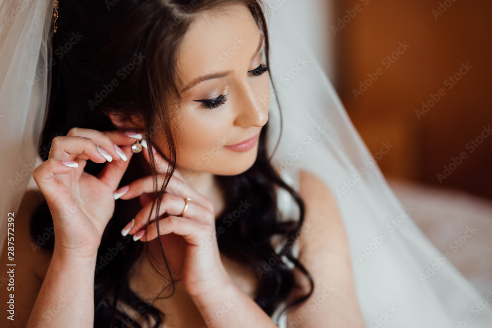Stylish happy bride corrects the earring, Beautiful earrings and hands of the bride. Wedding day. Happy woman bride in home prepared to wedding ceremony. Bride with elegant hairstyle and makeup