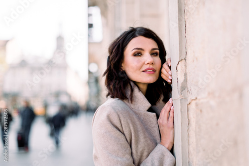 Closeup portrait of pretty girl with short dark hair smiling to camera in city on old building background. She wears light coat.