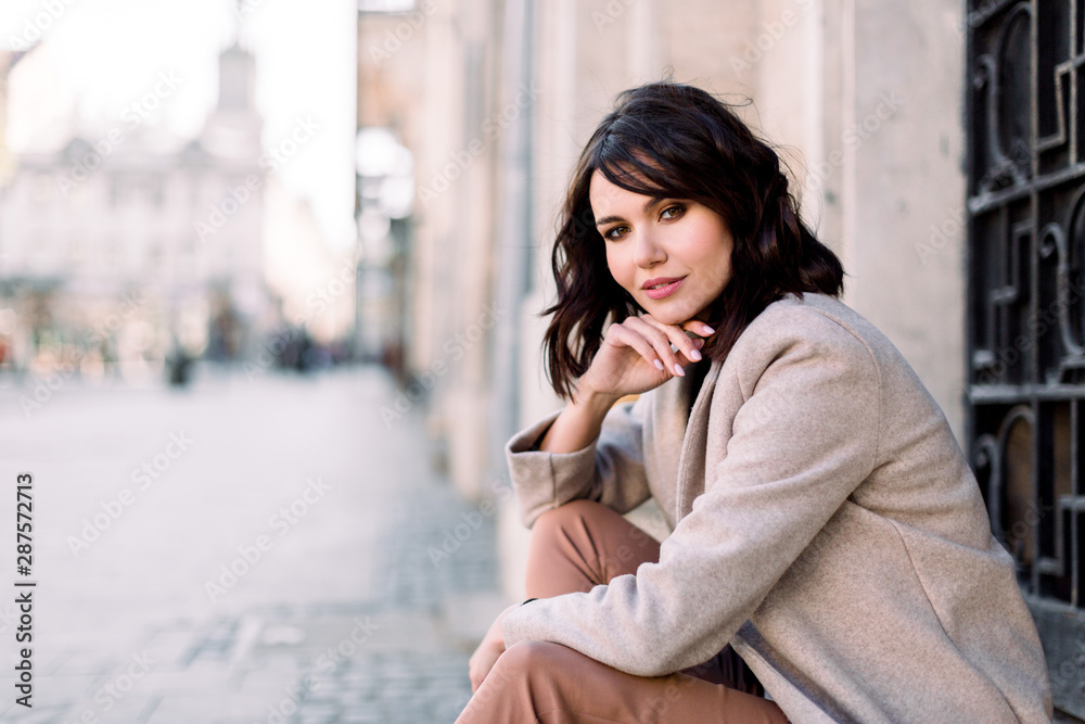 Portrait of young beautiful lady in brown pants and beige coat sitting on the stone stairs near the old building in city. City lifestyle. Copy space for text. Portrait of young beautiful lady in brown pants and beige coat sitting on the stone stairs near the old building in city. City lifestyle. Copy space for text.