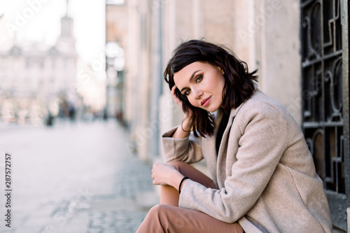 Close-up portrait of pretty girl with short brunette hair at street background wearing beige coat, make up. Smiling and sensual young woman posing while sitting on stairs © sofiko14