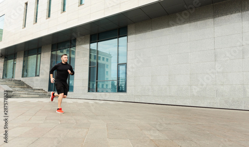 Serious millennial man jogging outdoors against urban building