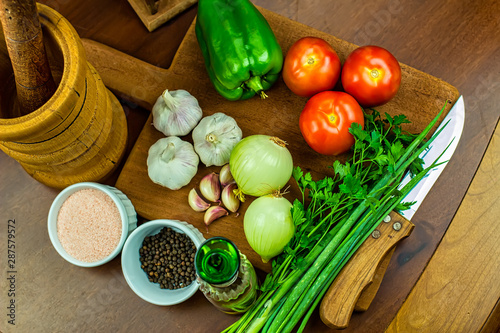 Garlic  onions  green peppers  tomatoes and parsley and chives on wooden cutting board with glass of olive oil  salt and black pepper in ramekins  wooden mortar and wooden chopping block in top view