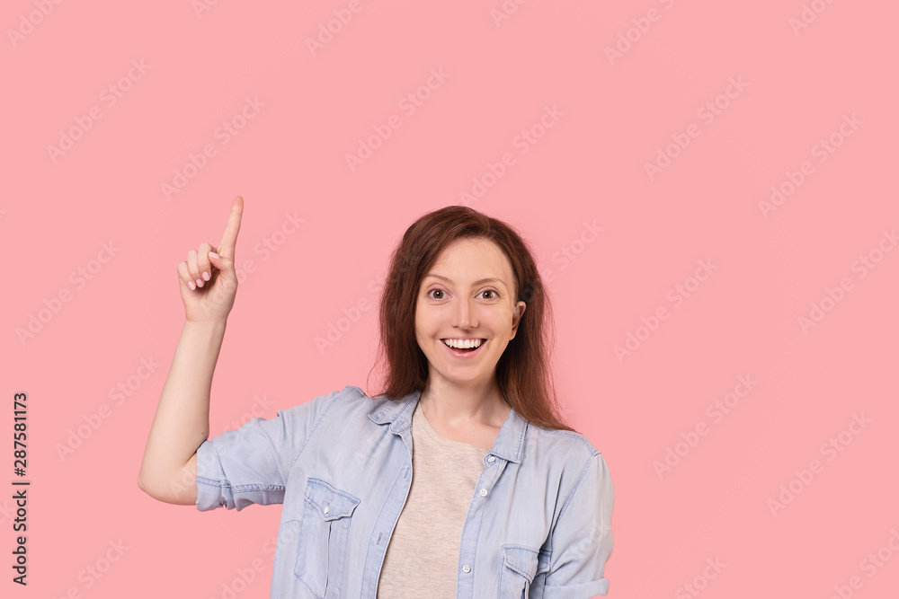 Emotional positive young woman in casual wear points finger up posing on pink background. Happy woman advertises personal offer and discount in the online store. Place for text Emotional positive young woman in casual wear points finger up posing on pink background. Happy woman advertises personal offer and discount in the online store. Place for text