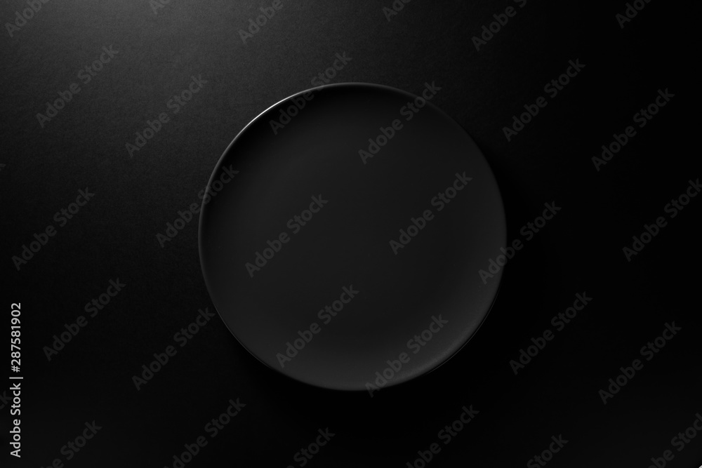 Empty round black plate on dark moody black background with copy space. Overhead view - obrazy, fototapety, plakaty 