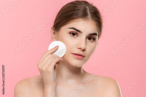 Beautiful model applyes cosmetic product to skin with white cotton sponge. Model looks at the camera isolated over pink background. Concept of beauty and health treatment.
