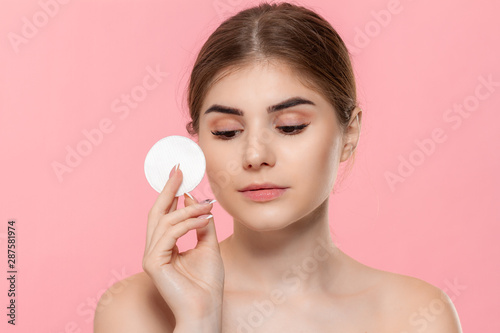 Beautiful model applyes cosmetic product to skin with white cotton sponge isolated over pink background. Concept of beauty and health treatment.
