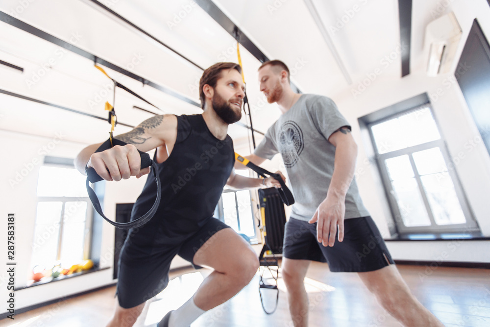 Fototapeta premium Male trainer teaches his student how to workout with suspended training system. Concept of strength training and work with the support of a coach