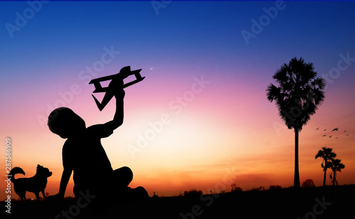 Silhouette of kid dreams as pilot sitting and holding airplane paper with friend running with wind turbine in sunset, imagination and freedom idea concept