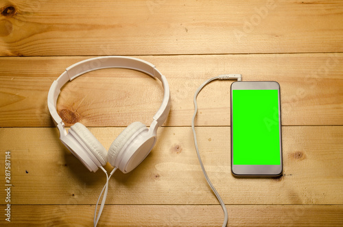 White headphones connected to phone on wooden table. Modern headphone with cable and gadget with green screen.