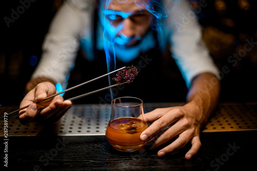 Professional bartender adding chilled melting caramel with twezzers to the cocktail with ice cubes under blue light