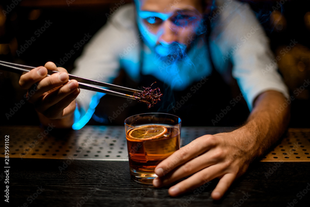 Professional bartender adding chilled brown caramel with twezzers to the cocktail with ice cubes under blue light Professional bartender adding chilled brown caramel with twezzers to the cocktail with ice cubes under blue light