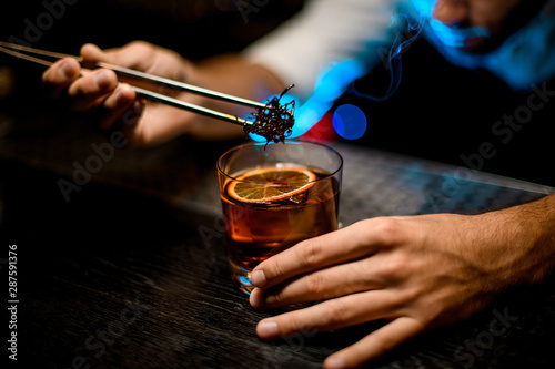 Professional bartender adding chilled melting caramel with twezzers to the cocktail with dried orange under blue light