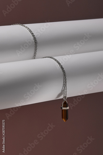 Close-up shot of a steely chainlet with a pendant in the form of screw with a fitted-in brown gemstone similar to a cat's-eye. The glazy gemstone is elongated and conical. 