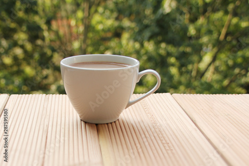 white cup with coffee on a light wooden table against a green summer garden, close-up, copy space