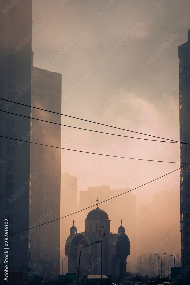 Φωτογραφία Beautiful scene of morning sunlight through the dense fog between high modern city buildings, and small christian orthodox church silhouette in the middle