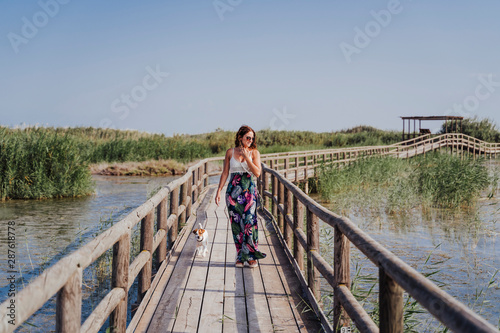 young beautiful woman walking on a wood pier in a natural park at sunset. Cute jack russell small dog looking at her owner.Tourism, love and travel concept