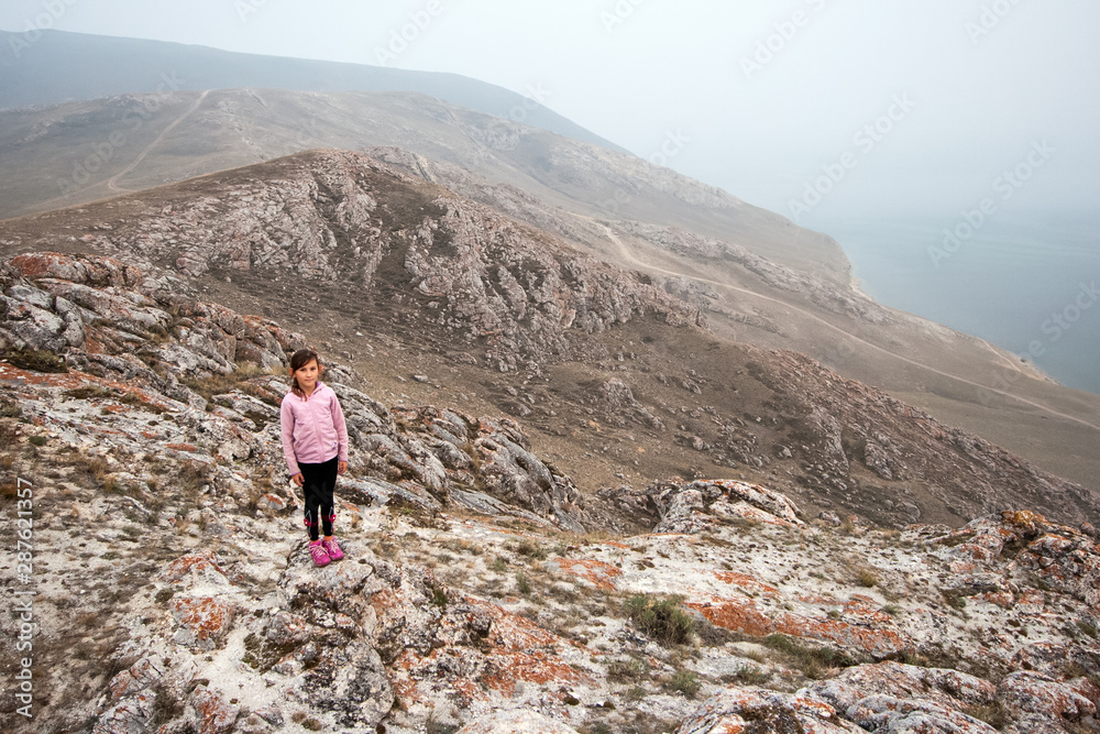 A young girl tourist stands on the edge of a high cliff on the shore of Lake Baikal. Girl looking at the camera. The cliffs are red covered with moss. The weather is foggy.