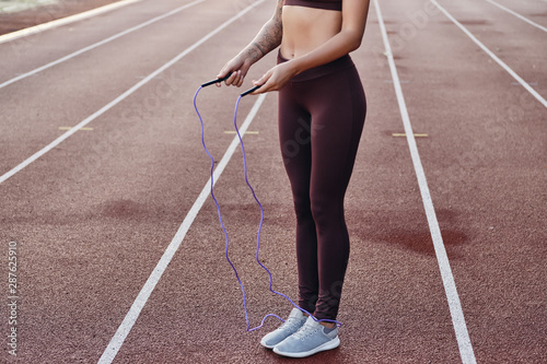Close up fitness girl in stylish sportswear using skipping rope while tranining on city stadium