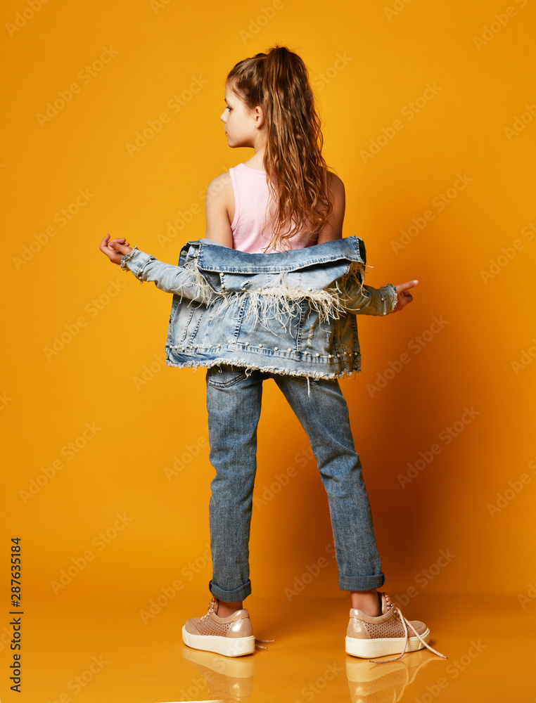 young teen girl model posing on a yellow background in jeans and a jacket,  with long hair gathered in a high tail, stands half a turn to the camera.  Stock Photo