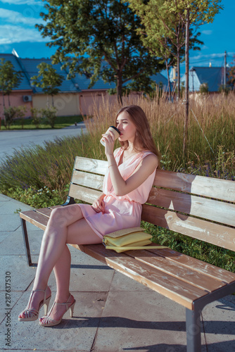 A girl sits on a bench in city park in summer, drinks tea from cup of coffee, handbag. Rest after work, lunch at lunch, break at work, background trees bushes, wooden bench. Pink heeled sandal dress.