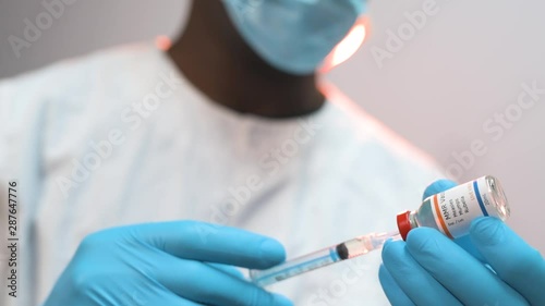 African American doctor drawing vaccine from vial into a syringe on a black background wearing white coat and blue gloves with mask photo