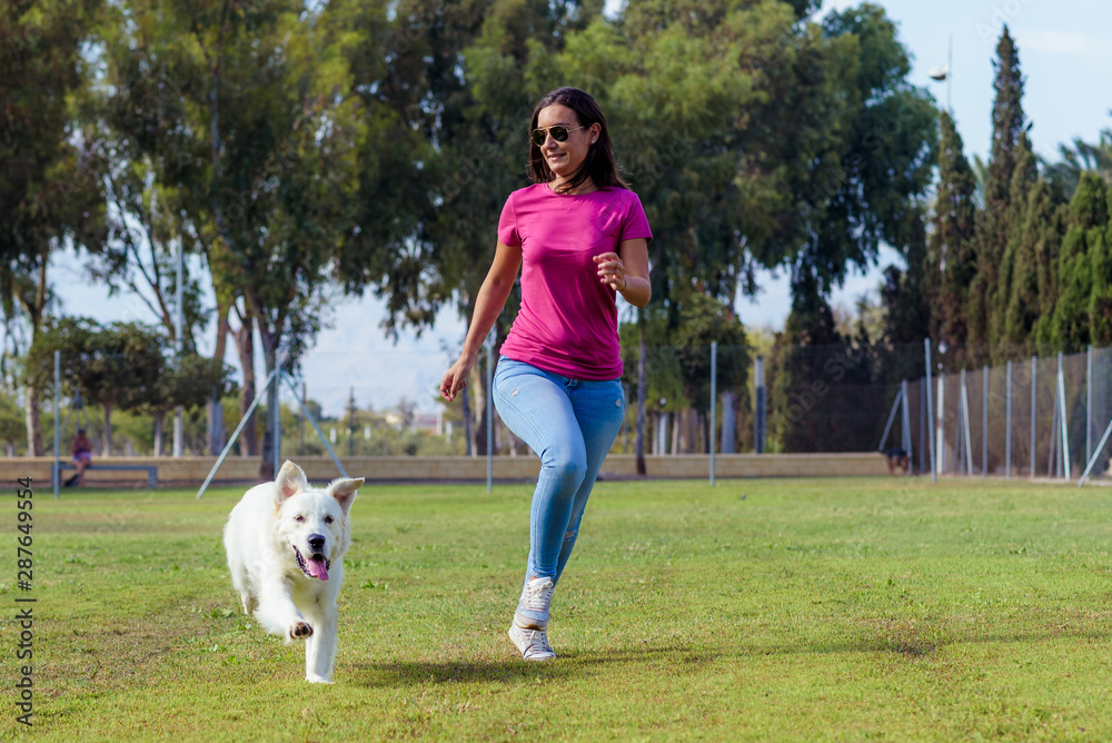 Sporty woman and dog running together in the park on summer sunset. Cheerful female athlete training and exercising outdoor with her pet. - obrazy, fototapety, plakaty 