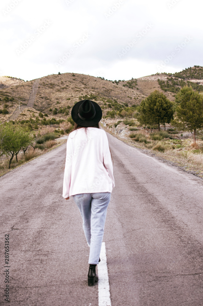 young woman with hat seen from behind walking alone on an empty road between mountains