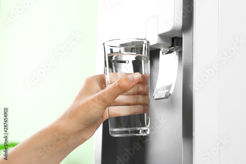 Woman filling glass from water cooler indoors, closeup. Refreshing drink