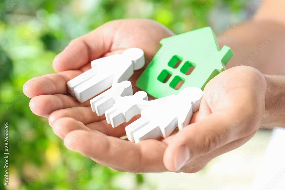 Young man holding wooden family figure and house model in his hands against blurred background, closeup Young man holding wooden family figure and house model in his hands against blurred background, closeup