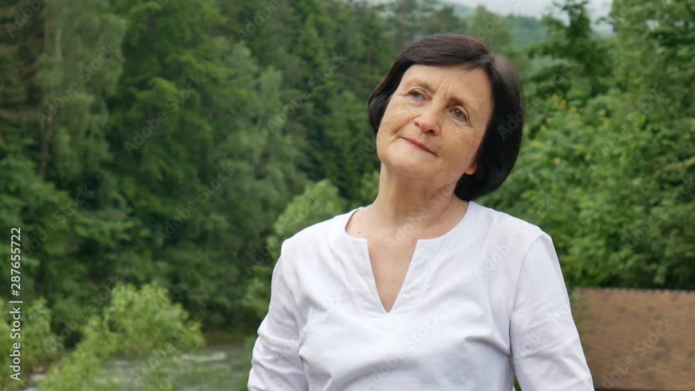 Outdoors portrait of an older woman with short dark hair and wrinkled face pointing at something during her trip to the mountains on mountain hill with green forest on background Outdoors portrait of an older woman with short dark hair and wrinkled face pointing at something during her trip to the mountains on mountain hill with green forest on background