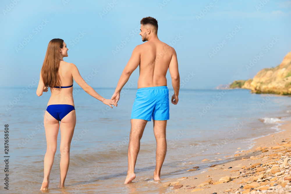 Young woman in bikini with her boyfriend walking on beach, space for text. Lovely couple