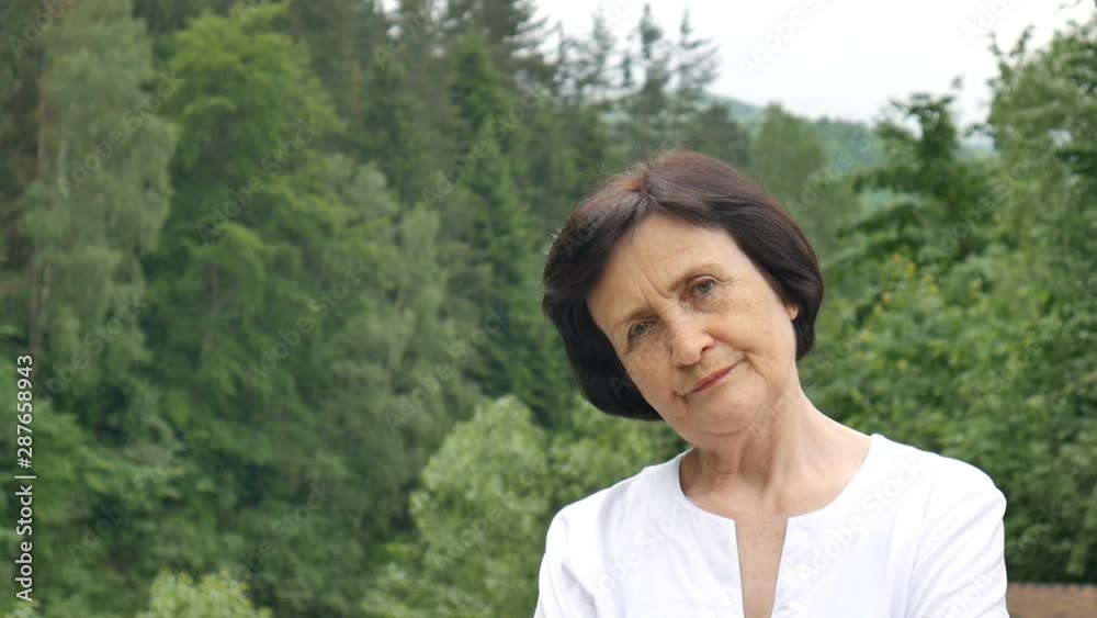 Female portrait of dissatisfied upset older woman with short dark hair and wrinkled face looking at the camera with disapproval outdoors on mountain hill with green forest on background Female portrait of dissatisfied upset older woman with short dark hair and wrinkled face looking at the camera with disapproval outdoors on mountain hill with green forest on background