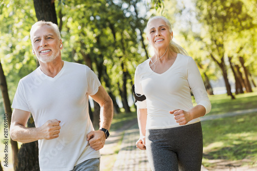 Portrait of elderly man and old woman in headphones jogging together outside in park