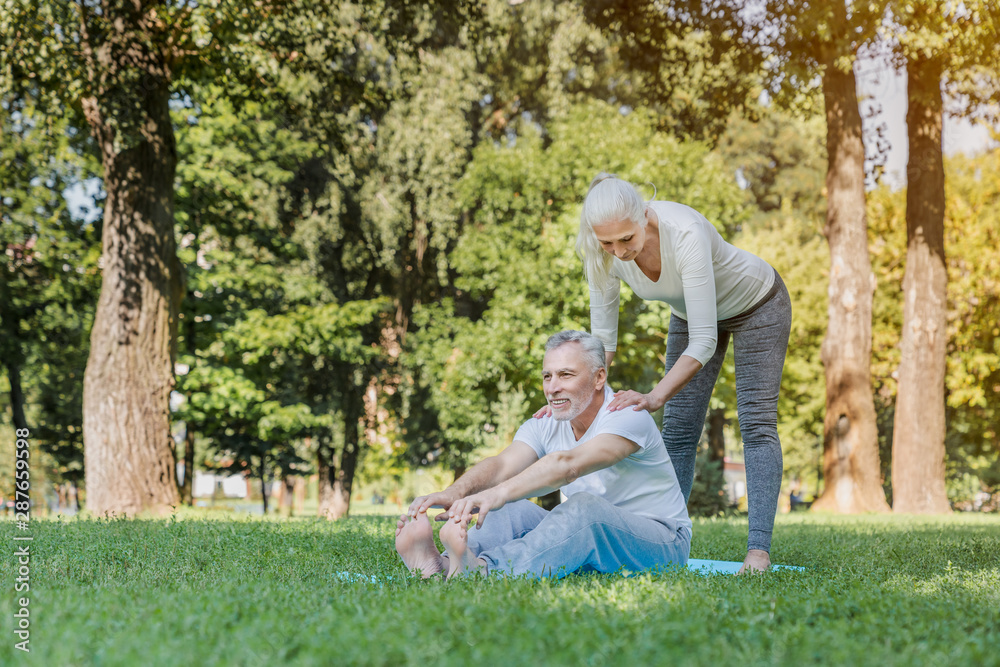 Fototapeta premium Senior woman helping to stretch mature man outdoors at park while doing workout and yoga exercices