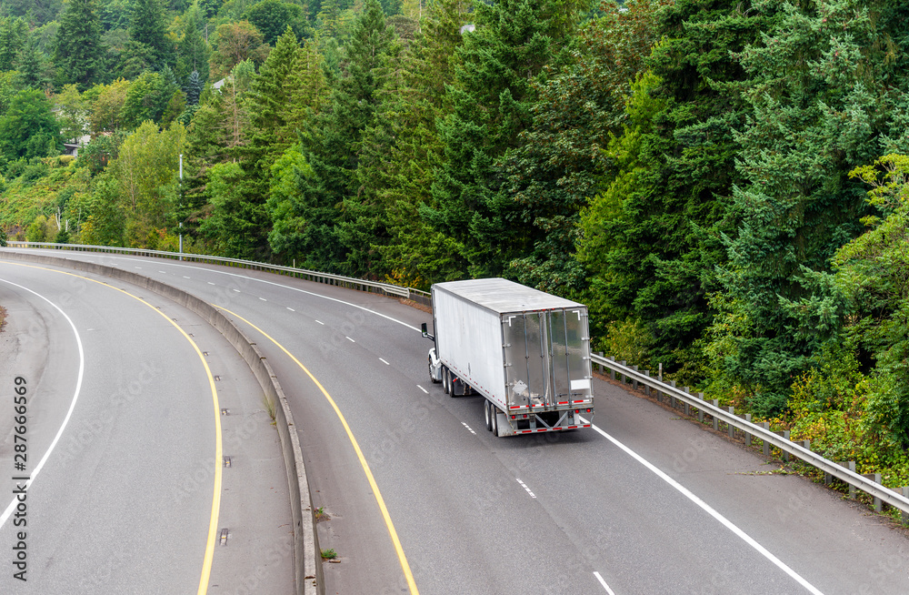 Big rig semi truck with semi trailer running on the turning divided road with green trees on the side Big rig semi truck with semi trailer running on the turning divided road with green trees on the side