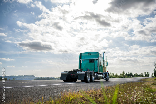 Green classic big rig semi truck tractor driving on the road with cloudy sky to warehouse for pick up the loaded trailer