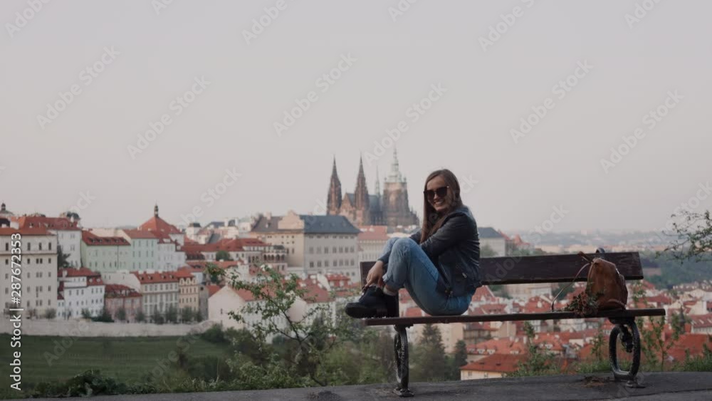 happy girl in sunglasses sits with legs on bench. There are traditional Czech roofs in background happy girl in sunglasses sits with legs on bench. There are traditional Czech roofs in background