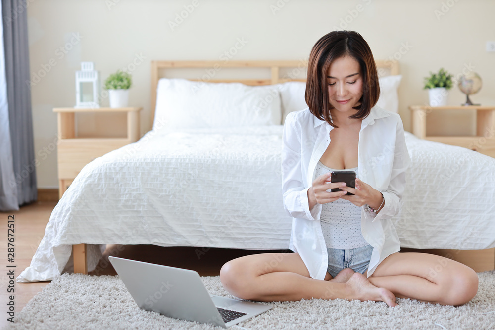 front view of adult freelance asian woman in white shirt working on computer and cell phone in bedroom with happy smiling face