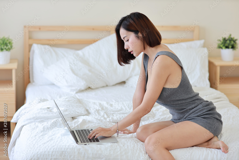 side view of adult freelance asian woman working on computer and cell phone on bed in bedroom side view of adult freelance asian woman working on computer and cell phone on bed in bedroom