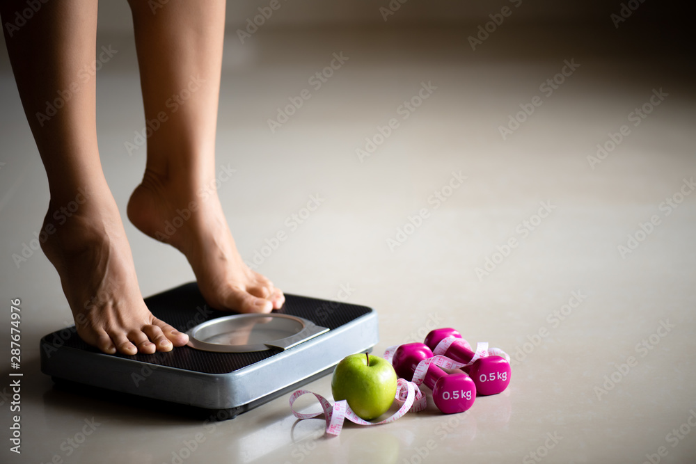 Female leg stepping on weigh scales with measuring tape and green apple. Healthy lifestyle, food and sport concept. - obrazy, fototapety, plakaty 