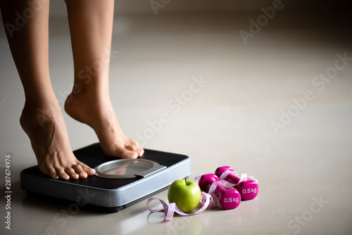 Female leg stepping on weigh scales with measuring tape and green apple. Healthy lifestyle, food and sport concept. photo